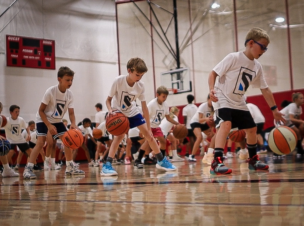 Young children bounce basketballs in unison while practicing a dribbling drill.