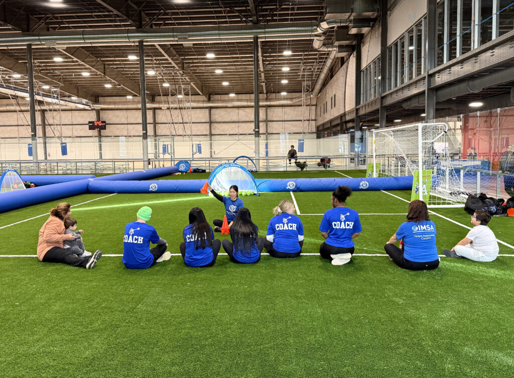 A group of Lil' Kickers soccer coaches sit on the turf field