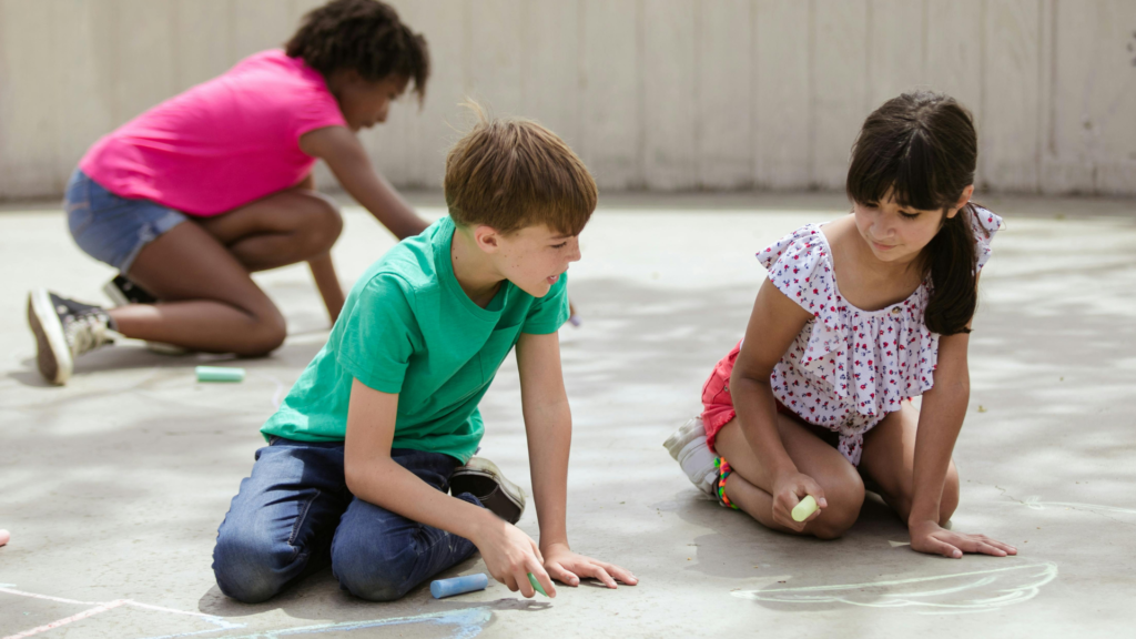Kids use chalk to draw on pavement outside during summer camp.
