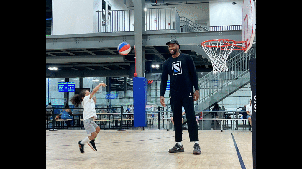 Coach smiling and supporting a 3-year-old basketball player shooting a ball into the hoop at Sawmill Sports Hub.