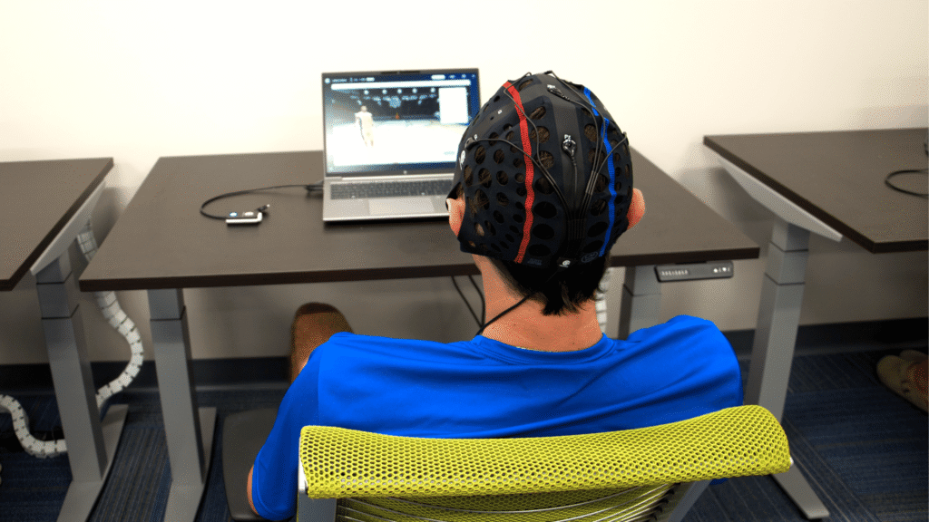 A young male athlete wearing a skull cap with visible wires connecting to a computer screen, focused intently while using the i-BrainTech system at the Synapse Brain Gym inside Sawmill Sports Hub.