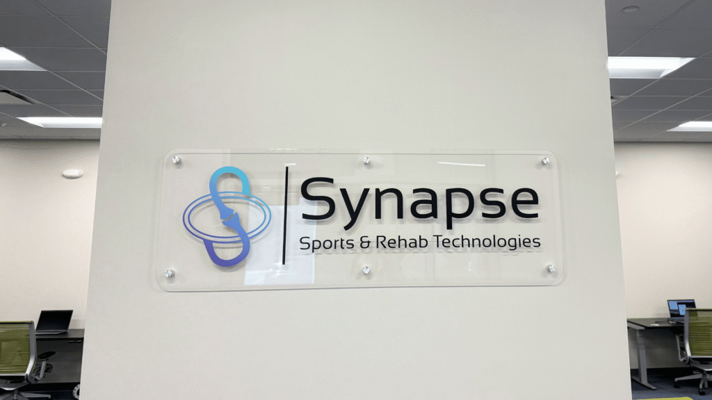 A close-up of the Synapse Sports & Rehab logo on a wall inside the Sawmill Sports Hub building, featuring the business name and an icon.