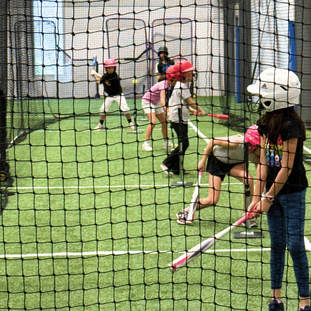 A modern batting cage facility, part of the Sawmill baseball and t-ball programs, with green turf and protective netting.