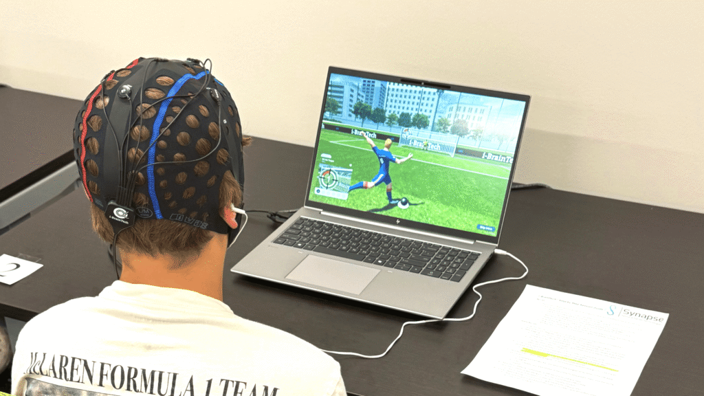 A young adolescent male athlete wearing a skull cap with visible wires connecting to a computer screen, focused intently while using the i-BrainTech system at the Synapse Brain Gym inside Sawmill Sports Hub.