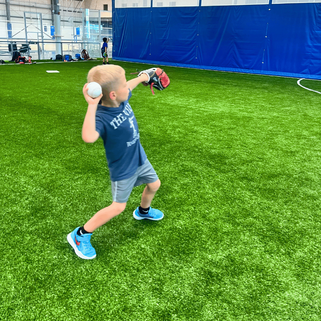 A young baseball player (aged 5) throws a baseball while holding his glove in his other hand at Sawmill Sports Hub's indoor baseball field.