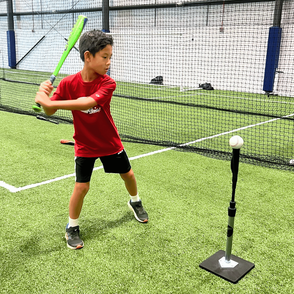 A young baseball player (aged 7) hits a baseball on a tee while holding a baseball bat at Sawmill Sports Hub's indoor baseball field.
